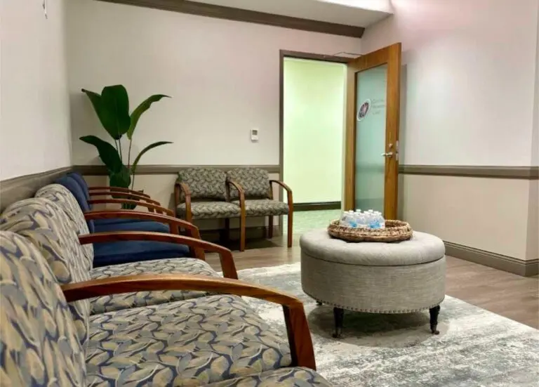 A waiting room with patterned cushioned chairs lining the walls. A circular ottoman sits in the center, topped with a basket of water bottles. A large plant is in the corner, and the door to the hallway is open.