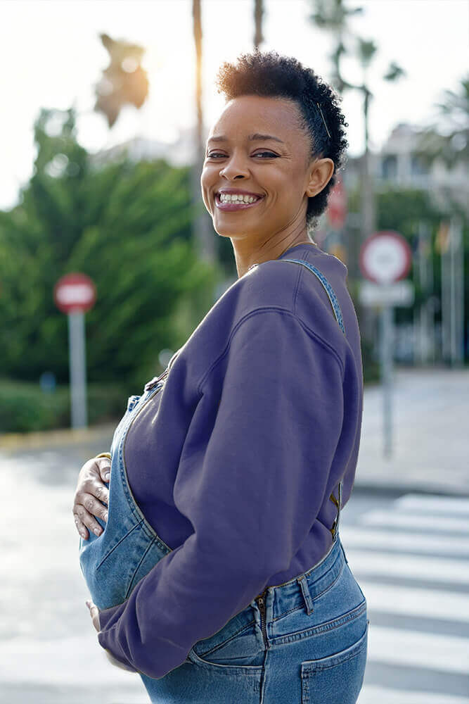 A smiling pregnant woman stands outdoors, gently holding her belly. She wears a purple sweatshirt and denim overalls. A pedestrian crossing and trees are visible in the background. Sunlight filters through the trees.
